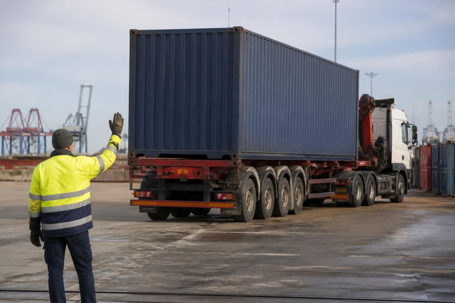 A standard 20ft shipping container sits on the back of a flatbed truck parked at a busy industrial port terminal