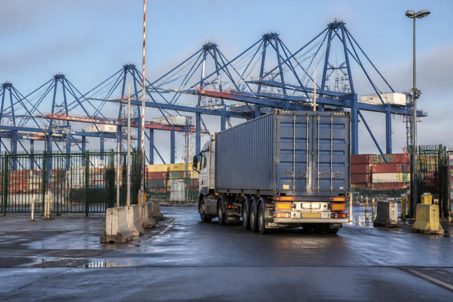 A truck driver stands beside a shipping container at a busy port terminal under a clear blue sky